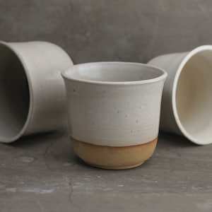 May include: Three white ceramic mugs, two with a simple rounded shape and one with a slightly flared lip. The mug with the flared lip has a brown band around the bottom.