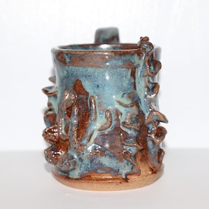 Puede incluir: Taza de cerámica hecha a mano con un diseño único. La taza presenta un esmalte azul y marrón con formas orgánicas texturizadas que adornan el exterior. Tiene un asa resistente y una base marrón claro. Una pieza funcional y artística.
