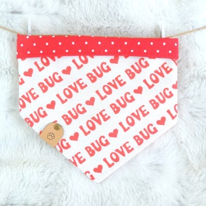 May include: White dog bandana with red polka dot trim and the words "Love Bug" repeated in red. The bandana has a brown leather patch with a paw print.
