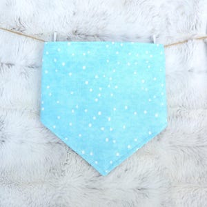 May include: A light blue, triangular bandana with white polka dots, hanging from a string. The bandana is made of soft fabric and is likely for a pet. The background is a fluffy, white surface.