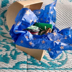 May include: A heart-shaped box filled with blue tissue paper with gold stars. The box contains a green and brown candle, a small jar of green and brown wax, and a small jar of brown wax.