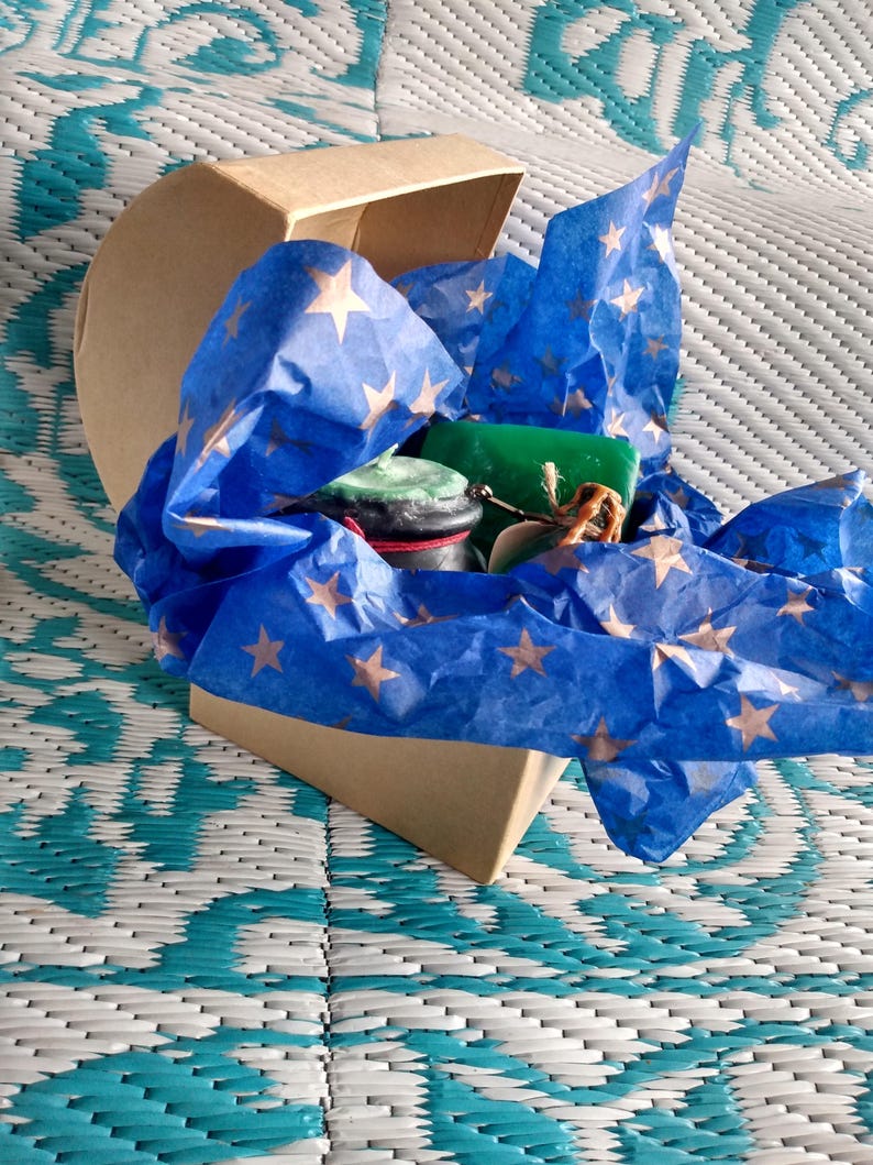 May include: A heart-shaped box filled with blue tissue paper with gold stars. The box contains a green and brown candle, a small jar of green and brown wax, and a small jar of brown wax.