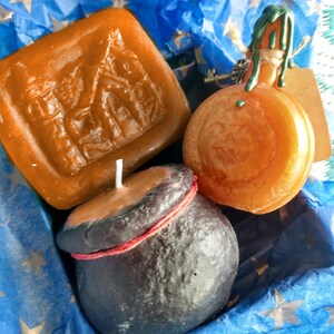 May include: Three candles in a blue and gold star patterned gift box. The candles are shaped like a house, a cauldron, and a pumpkin. The house candle is brown, the cauldron is black, and the pumpkin is orange.