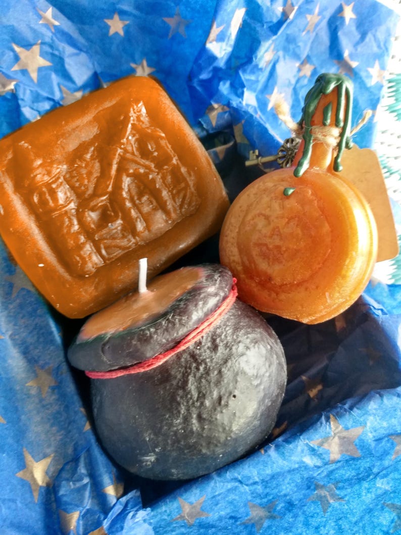 May include: Three candles in a blue and gold star patterned gift box. The candles are shaped like a house, a cauldron, and a pumpkin. The house candle is brown, the cauldron is black, and the pumpkin is orange.