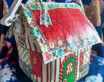 May include: A whimsical gingerbread house-shaped handbag with a white handle. The bag features a red roof, green door, and colorful candy and gingerbread man decorations. The bag is made of a glossy material and has a white handle.