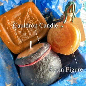 May include: A gift set of three handmade candles and a resin figure. The candles are shaped like a cauldron, a pumpkin, and a bar of soap. The resin figure is a cauldron with a red rope around the top. The candles are all orange and the resin figure is black.