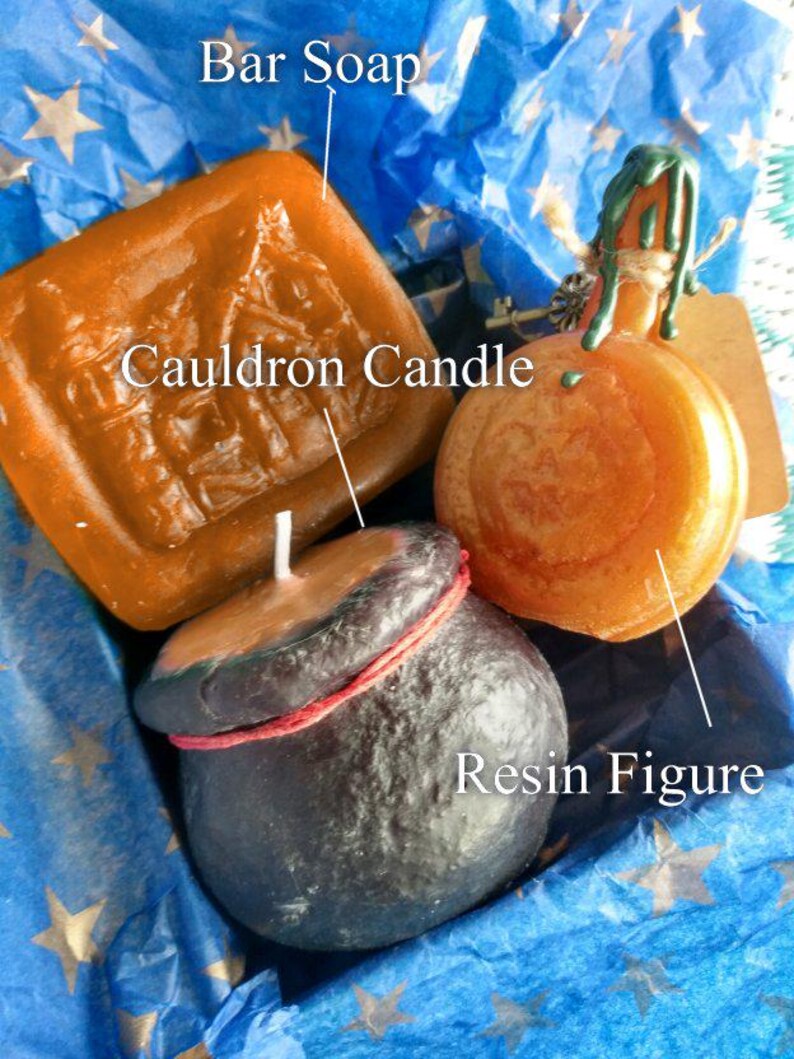 May include: A gift set of three handmade candles and a resin figure. The candles are shaped like a cauldron, a pumpkin, and a bar of soap. The resin figure is a cauldron with a red rope around the top. The candles are all orange and the resin figure is black.