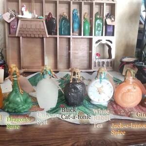 May include: Assortment of small glass bottles with various colored liquids and decorative stoppers. The bottles are labeled with names like "Buttered Beere", "Dragon's Bane", "Unicorn Spit", "Black Cat-a-tonic", "White Rabbit Tea", "Jack-o-lantern Spice", and "Gingerbread".