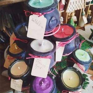 May include: A collection of black cauldron-shaped candles with various scents, including "Wildfire" and "Unicorn Spit". The candles are arranged on a tiered stand and have different colored waxes, including green, red, white, yellow, and purple.