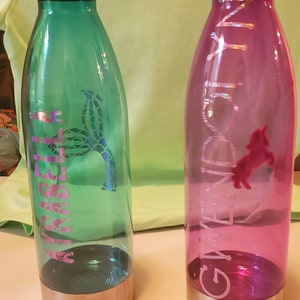 May include: Two reusable water bottles with personalized names. The bottle on the left is green with a mermaid design and the name "Gabrielle" on it. The bottle on the right is pink with a unicorn design and the name "Gwenlyn" on it.