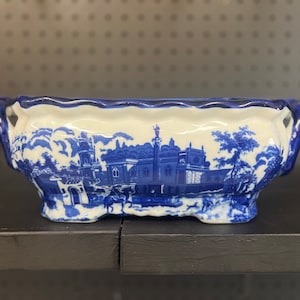 May include: A blue and white porcelain serving dish with a scalloped edge and a detailed design of a European village scene.