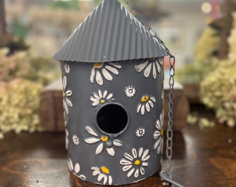 May include: A gray metal birdhouse with a white daisy pattern and a small black bird on top. The birdhouse has a hole for birds to enter and a chain for hanging.
