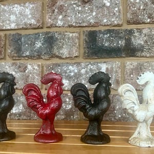 Op de afbeelding: Vier gietijzeren haanfiguren in verschillende kleuren: zwart, rood, zwart en wit. De hanen staan op een houten oppervlak.