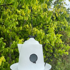 May include: White metal birdhouse shaped like a teapot with a black bird finial and a black rimmed saucer. The birdhouse has a wavy texture and a round hole for birds to enter.