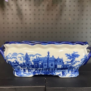 May include: A blue and white ceramic planter with a scalloped rim and two handles. The planter features a blue and white design of a cityscape.