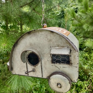 May include: A metal birdhouse shaped like a vintage camper trailer, painted in a distressed off-white color. Features a round door, a window with wire mesh, and a wheel. The top has the number "50" in a yellow rectangle. Suspended by a chain.