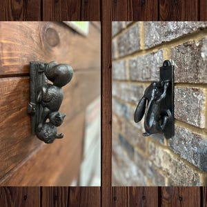 Könnte beinhalten: Zwei Türklopfer aus schwarzem Gusseisen. Einer zeigt ein Eichhörnchen, das einen Pfosten erklimmt, der andere ein Kaninchen. Beide sind auf rechteckigen Rückplatten montiert und an einer Holztür und einer Backsteinmauer befestigt.