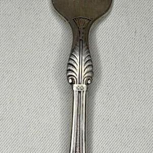 May include: A silver-toned metal fork featuring four tines and an ornate handle. The handle displays a vertical fluted design near the neck, a central beaded line, and a decorative floral motif at the base with an engraved letter L.