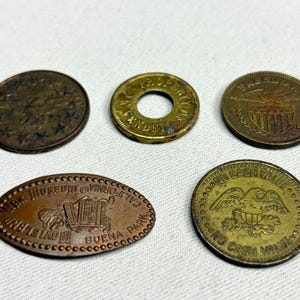 May include: A collection of five metal tokens and coins. One is a dark circular coin with stars, one is a brass-colored washer-style token, one is a circular token with a shield and eagle, one is an oval token marked Wax Museum Buena Park, and one is a gold-toned token with an eagle and the text No Cash Value.
