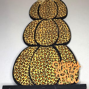 May include: A decorative wooden sign shaped like a stack of three pumpkins with a yellow and black leopard print pattern. The sign features a raffia bow at the top and an orange cutout script text that reads Happy Fall at the bottom.