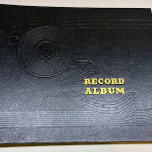 May include: A black record album with a textured cover featuring an embossed design of a vinyl record and musical staff lines. The words RECORD ALBUM are printed in gold lettering on the front center of the cover.