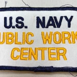 May include: A rectangular embroidered patch with a white background and a dark blue border. The text U.S. NAVY is stitched in dark blue, while the words PUBLIC WORKS CENTER are stitched in yellow thread below it.