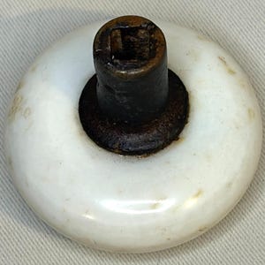 Puede incluir: Un pomo de puerta redondo de porcelana blanca con un soporte central de metal marrón oscuro y una abertura cuadrada. La superficie blanca muestra signos de desgaste y antigüedad, mientras que el metal tiene un acabado oxidado.