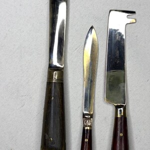 May include: A set of three metal utensils with dark wooden handles and ornate silver-toned metal ends. The left utensil is a long knife, the center is a small pointed knife, and the right is a flat-edged tool with a notched top.