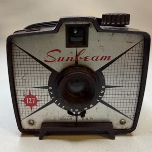 May include: A vintage Sunbeam box camera with a silver faceplate featuring a grid pattern, a red 127 star logo, and a Galtex 60 mm lens. The camera body is dark brown with a top-mounted winding knob and a front-facing viewfinder window.