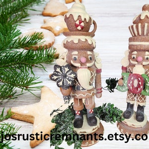 May include: Two gingerbread-style nutcrackers with white frosting accents and holding a snowflake and a wreath that says "JOY". The nutcrackers are brown and beige with red and white details. The background includes star-shaped cookies and evergreen branches.