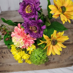 May include: A bouquet of artificial flowers in a wooden crate. The bouquet includes purple, pink, yellow, and green flowers.