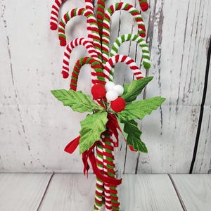 May include: A festive Christmas decoration featuring a bouquet of red and white striped candy canes with green and white stripes, tied together with a red ribbon and accented with green leaves and white pom-poms.
