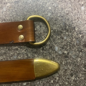 Pu&ograve; includere: Primo piano di una cintura in pelle marrone con fibbia e rivetti in ottone. La cintura ha un anello a D color ottone e una punta color ottone. La pelle sembra usurata.