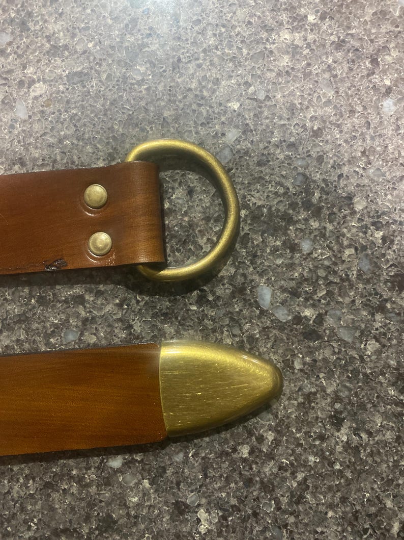 Pu&ograve; includere: Primo piano di una cintura in pelle marrone con fibbia e rivetti in ottone. La cintura ha un anello a D color ottone e una punta color ottone. La pelle sembra usurata.