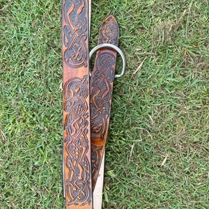 Pu&ograve; includere: Una cintura in pelle marrone con intricate incisioni di nodi celtici. La cintura presenta una fibbia color argento e un design dettagliato su tutta la sua lunghezza. La cintura &egrave; appoggiata sull'erba verde.