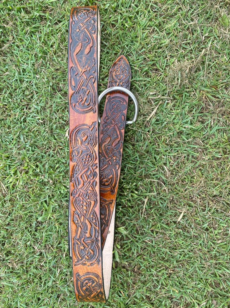 Pu&ograve; includere: Una cintura in pelle marrone con intricate incisioni di nodi celtici. La cintura presenta una fibbia color argento e un design dettagliato su tutta la sua lunghezza. La cintura &egrave; appoggiata sull'erba verde.