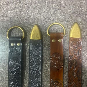 Pu&ograve; includere: Quattro cinture in pelle con finiture in ottone. Due sono nere, una marrone e una marrone rossiccio. Ogni cintura presenta un diverso motivo a nodo celtico. Le cinture hanno anelli a D in ottone e punte in ottone.
