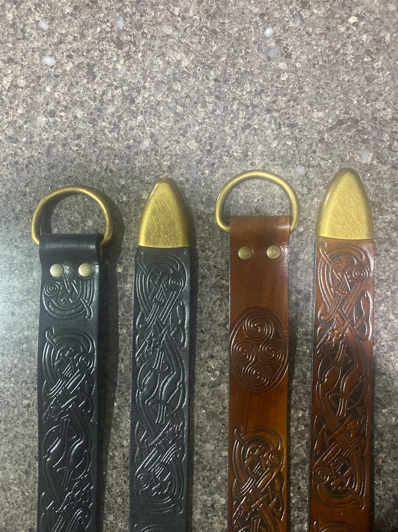 Pu&ograve; includere: Quattro cinture in pelle con finiture in ottone. Due sono nere, una marrone e una marrone rossiccio. Ogni cintura presenta un diverso motivo a nodo celtico. Le cinture hanno anelli a D in ottone e punte in ottone.