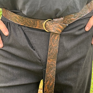 Pu&ograve; includere: Cintura in pelle marrone con un motivo a nodo celtico in rilievo. La cintura presenta una fibbia circolare color ottone e viene indossata sopra pantaloni neri. La pelle ha un aspetto invecchiato.