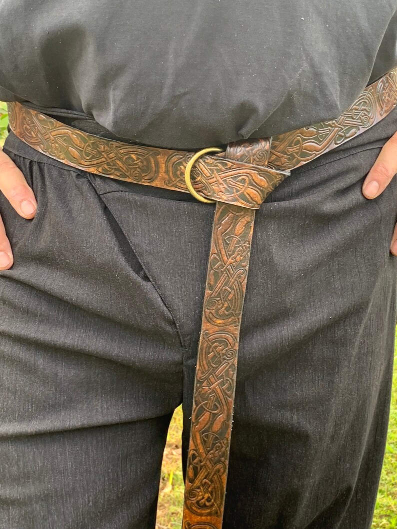 Pu&ograve; includere: Cintura in pelle marrone con un motivo a nodo celtico in rilievo. La cintura presenta una fibbia circolare color ottone e viene indossata sopra pantaloni neri. La pelle ha un aspetto invecchiato.