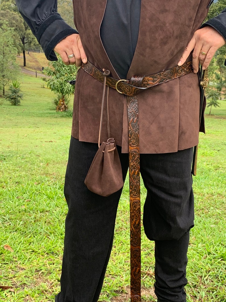 Pu&ograve; includere: Una cintura in pelle marrone con intricati disegni intagliati, una grande fibbia in ottone e una piccola borsa. La cintura &egrave; indossata con un gilet marrone e pantaloni neri, suggerendo un tema medievale o fantasy.