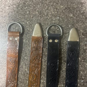 Pu&ograve; includere: Quattro cinture in pelle con dettagli metallici. Le cinture sono in tonalit&agrave; marrone e nera, ognuna con un motivo a nodo celtico. Le cinture hanno un anello e una punta in metallo. Sono esposte su una superficie grigia strutturata.