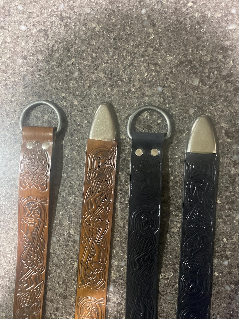 Pu&ograve; includere: Quattro cinture in pelle con dettagli metallici. Le cinture sono in tonalit&agrave; marrone e nera, ognuna con un motivo a nodo celtico. Le cinture hanno un anello e una punta in metallo. Sono esposte su una superficie grigia strutturata.