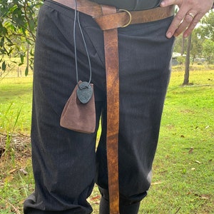 Pu&ograve; includere: Una cintura in pelle marrone con fibbia ad anello in ottone e un'estremit&agrave; lunga e affusolata. Una piccola borsa marrone con un ciondolo a nodo celtico pende dalla cintura. La persona indossa pantaloni neri.