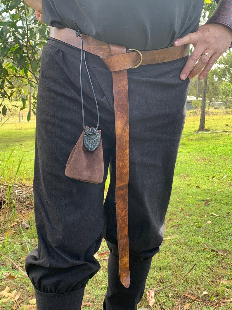 Pu&ograve; includere: Una cintura in pelle marrone con fibbia ad anello in ottone e un'estremit&agrave; lunga e affusolata. Una piccola borsa marrone con un ciondolo a nodo celtico pende dalla cintura. La persona indossa pantaloni neri.
