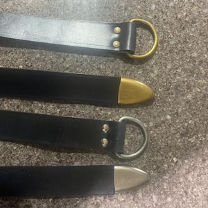 Pu&ograve; includere: Quattro cinture in pelle nera con dettagli metallici. La cintura superiore ha un anello a D color oro, la seconda ha una punta color oro, la terza ha un anello a D argentato e l'inferiore ha una punta argentata. Le cinture sono su una superficie strutturata.