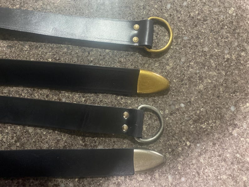 Pu&ograve; includere: Quattro cinture in pelle nera con dettagli metallici. La cintura superiore ha un anello a D color oro, la seconda ha una punta color oro, la terza ha un anello a D argentato e l'inferiore ha una punta argentata. Le cinture sono su una superficie strutturata.