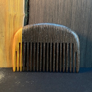 May include: A wooden comb with a dark brown and orange wood grain. The comb has a rounded top and 15 teeth.