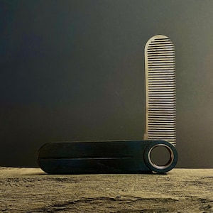 May include: A black folding comb with a silver metal comb section. The comb is closed and sitting on a piece of wood.