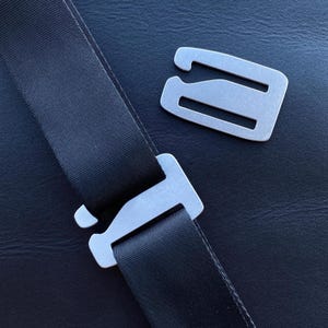 May include: A close-up shot of a black seatbelt with a silver metal buckle and a separate silver metal clasp. The seatbelt is made of a durable, woven material. The background is a dark, textured surface.